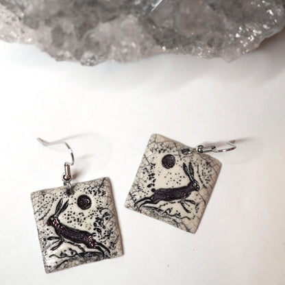 Black and white hand-made ceramic square dangle earrings featuring a scene of a wild rabbit running beneath a full moon