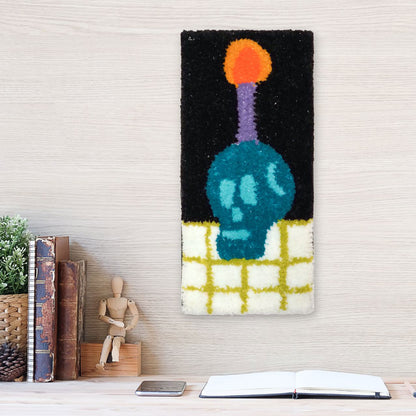 Hand-tufted wall hanging featuring a teal blue-colored skull on a white and green checked surface with a purple candle coming out of the top of its head