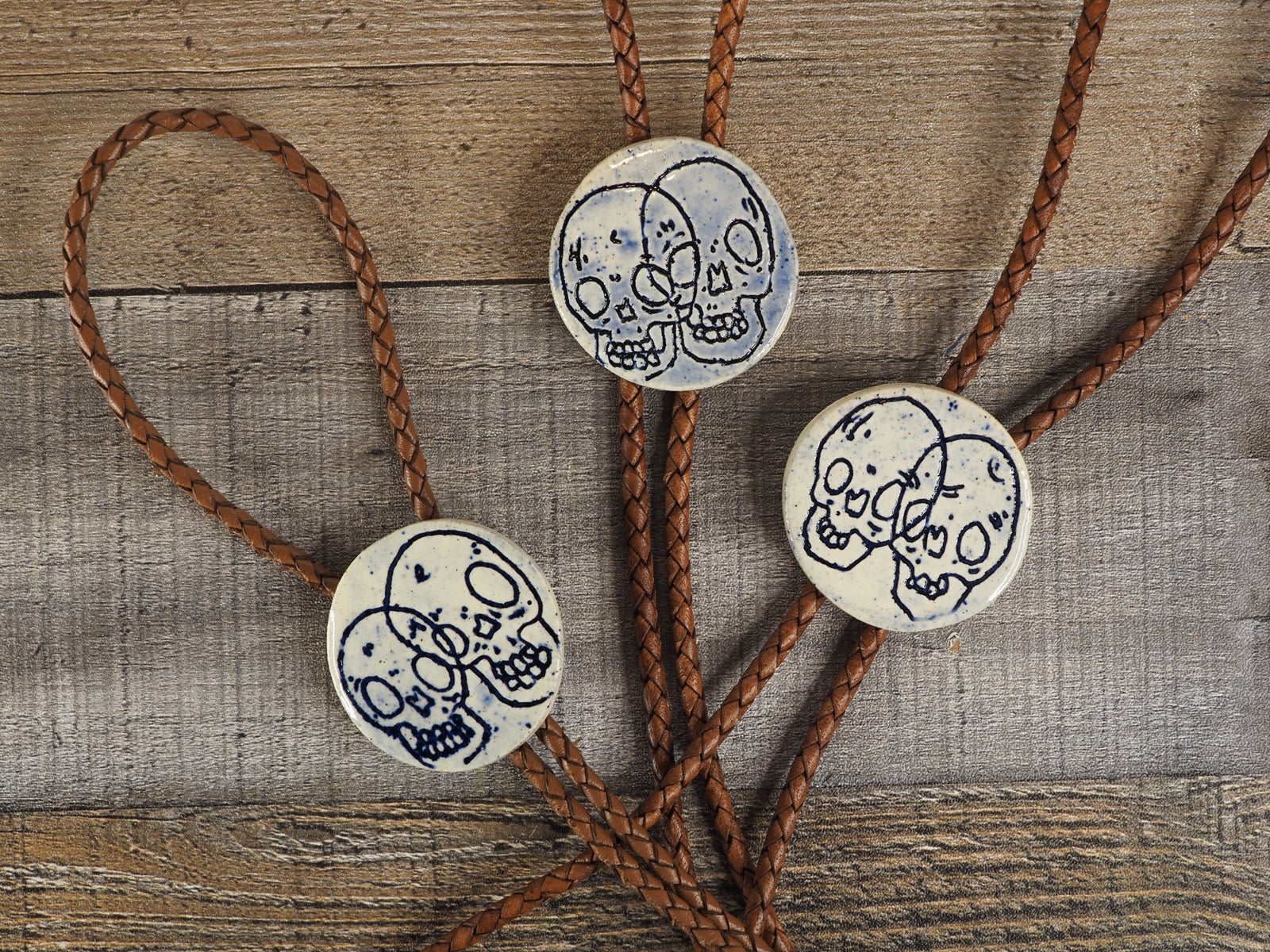 Skull illustration is carved and hand-painted onto white stoneware pendant with cobalt blue inlay, bolo tie hung on leather cords - 3 are shown together
